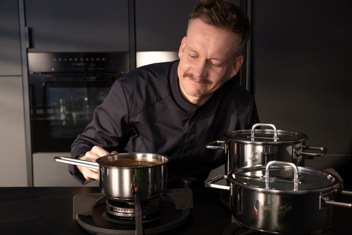 A chef in a modern kitchen enjoys a sauce simmering in a stainless steel Cooking Pot, with Fissler Viseo® Pro pots nearby.