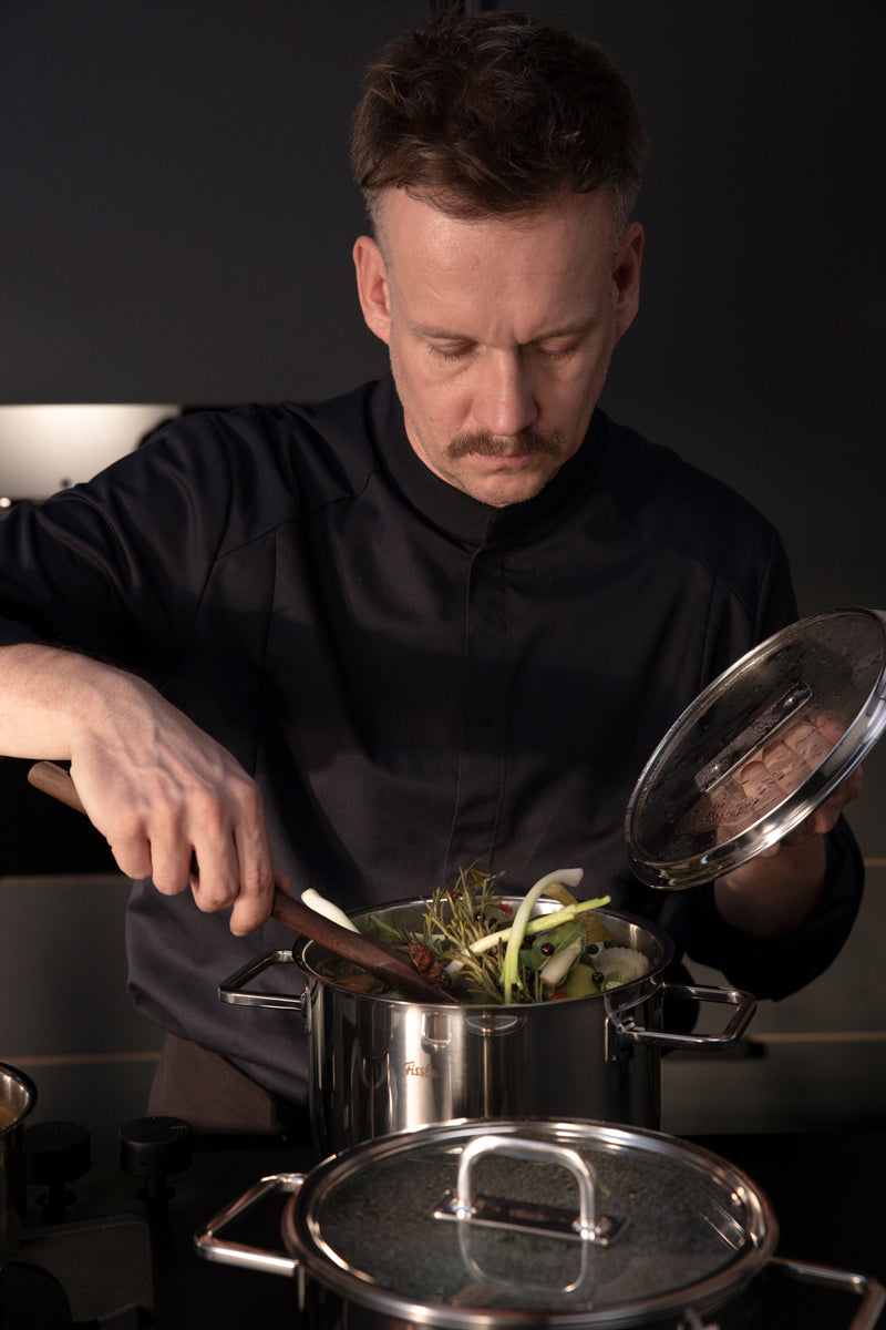 A man cooks with the Fissler Viseo® Pro Cooking Pot, holds the Lid, and stirs vegetables - precision and quality Made in Germany.