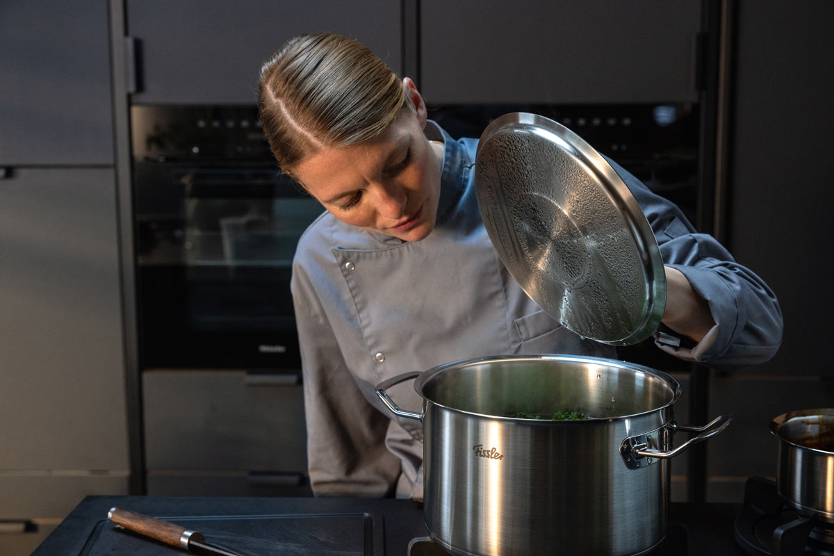 A chef in a gray uniform lifts the lid from a Fissler Original-Profi Collection® 5-piece stainless steel pot set on the stove, showcasing German-made precision and innovative design while preparing a meal in a sleek, modern kitchen.