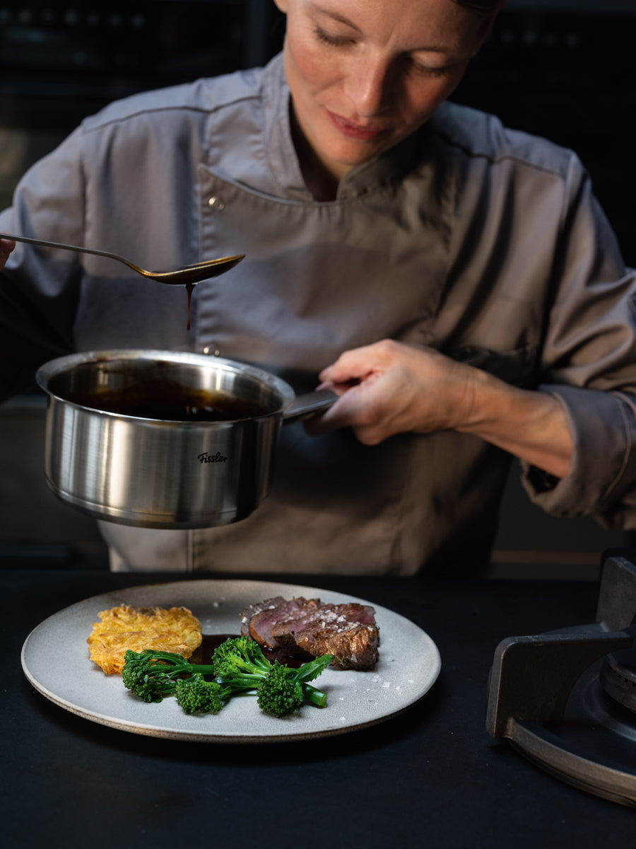Ein Koch in grauer Uniform demonstriert die außergewöhnliche Leistungsfähigkeit des 5-teiligen Sets der Original-Profi Collection® von Fissler – bestehend aus Töpfen, einem Auflauf, einem Stieltopf und einer Servierpfanne, die fachmännisch aus hochwertigem Edelstahl 18/10 „Made in Germany“ gefertigt wurden. Mit dem präzise gefertigten Kochgeschirr gießt der Koch sorgfältig Soße über ein kunstvoll angerichtetes Gericht mit Steak, Broccolini und Kartoffeln und unterstreicht damit Fisslers Engagement für