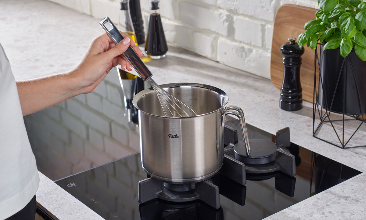 A person uses a wire whisk to stir inside a Fissler Original-Profi Collection® Milchtopf 16cm, precision-engineered for durability, on a gas stovetop.