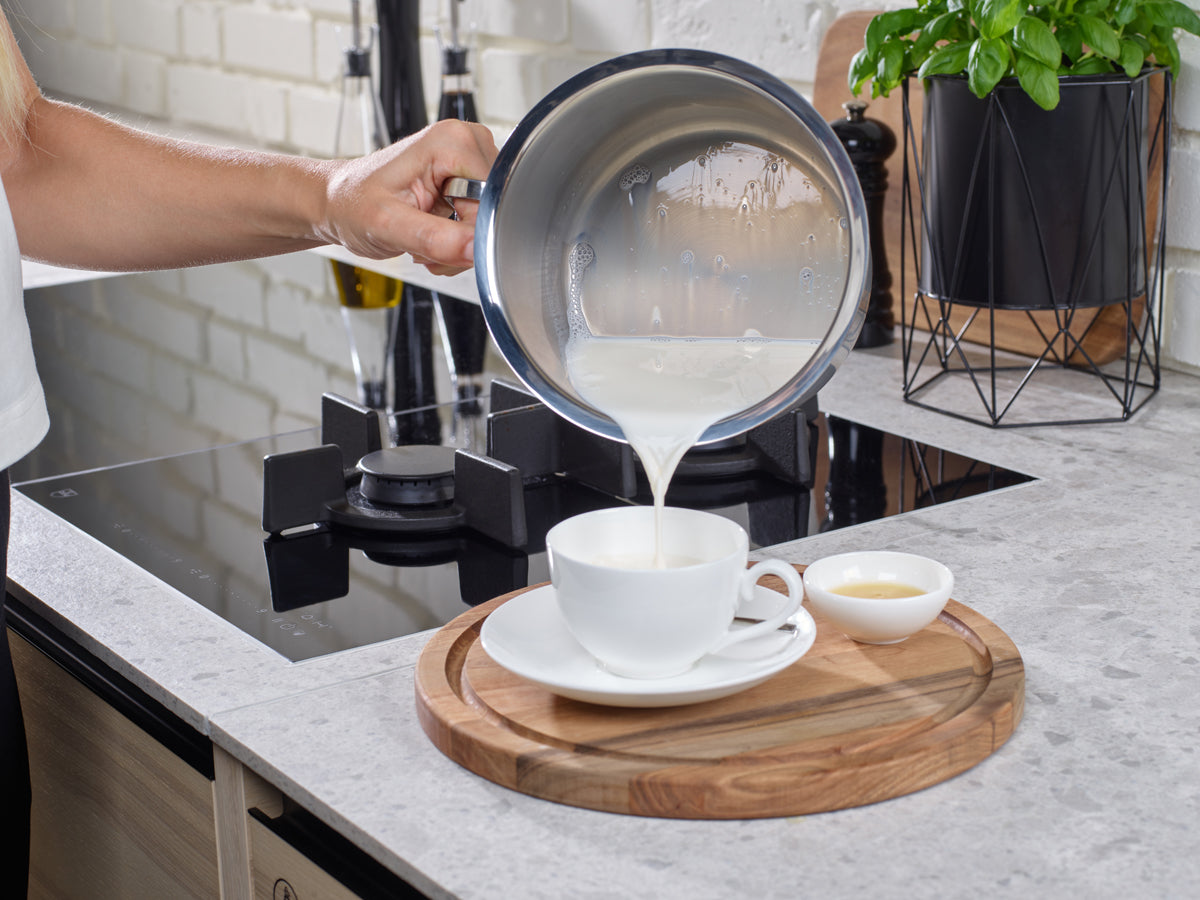 A hand pours milk from a Fissler Original-Profi Collection® 16cm milk pot, featuring German-engineered precision, into a white teacup resting on a wooden board.