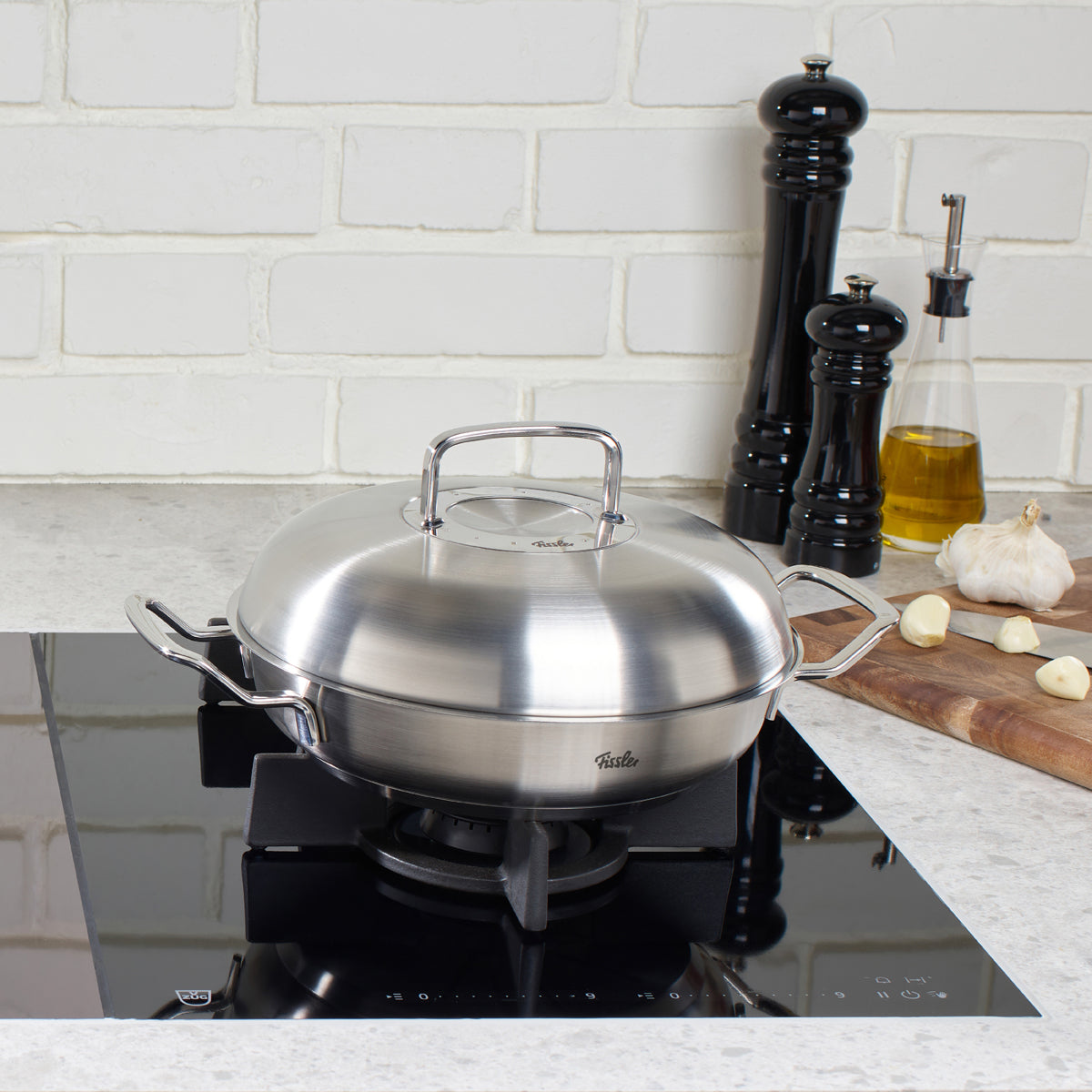 Fissler Original-Profi Collection® Serving Pan with Novogrill® Lid on a gas stove, surrounded by salt, pepper and olive oil.