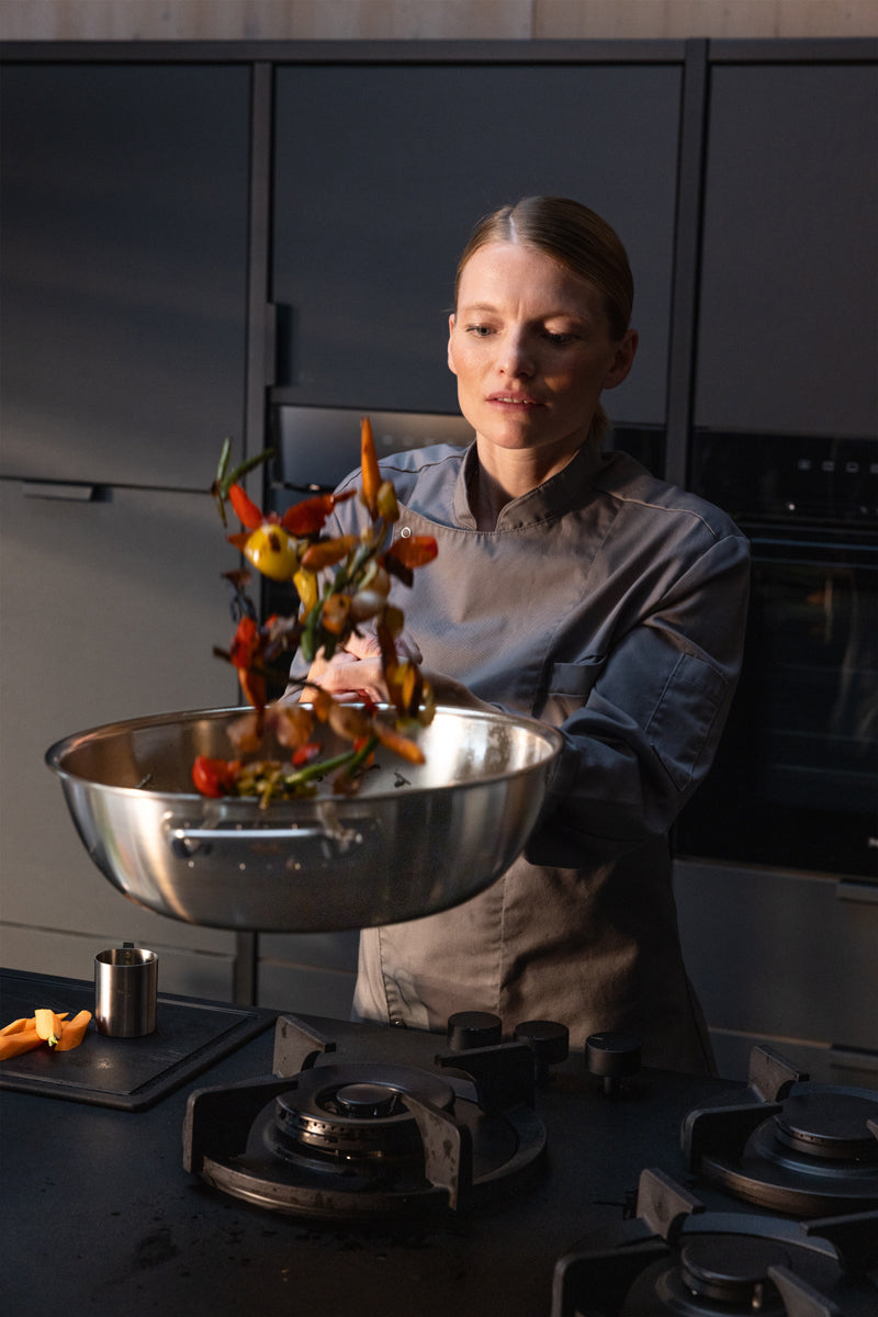 A chef in a gray uniform is vigorously sautéing colorful vegetables in a Fissler stainless steel pan over a stove in a sleek, modern kitchen.