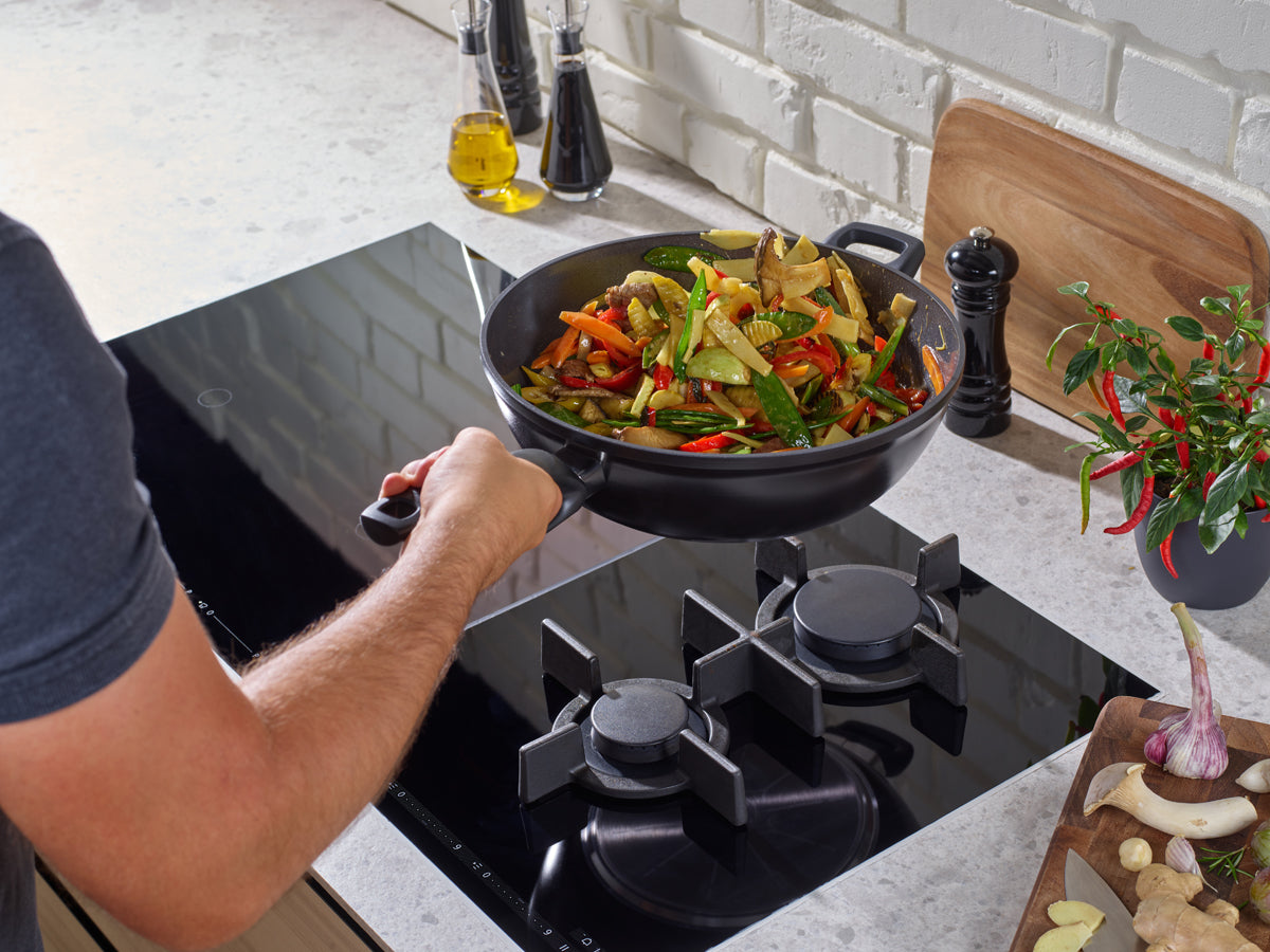A vegetable stir-fry is prepared in a Fissler Ceratal® wok in orbit black; spices and fresh ingredients are laid out on a simple, modern counter.