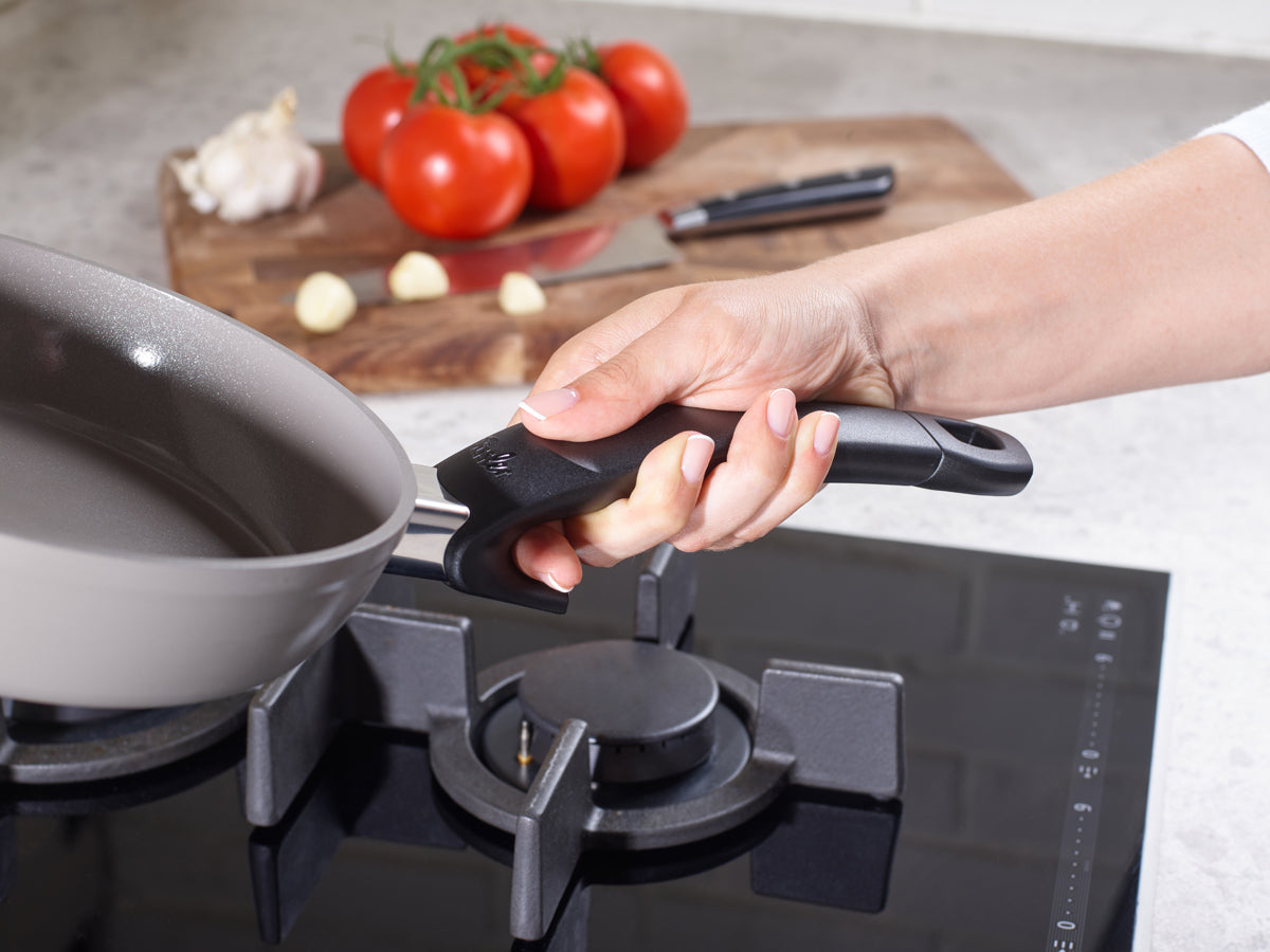 A black handle is attached to a Fissler Ceratal® Frying Pan on a gas stove, with fresh ingredients and quality German-made craftsmanship.