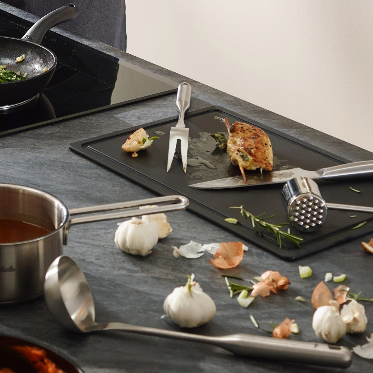 Cooked chicken on a board with Fissler Essential® meat fork, tenderizer; Fissler Cooking Pot and ladle on the stove; modern kitchen scene.