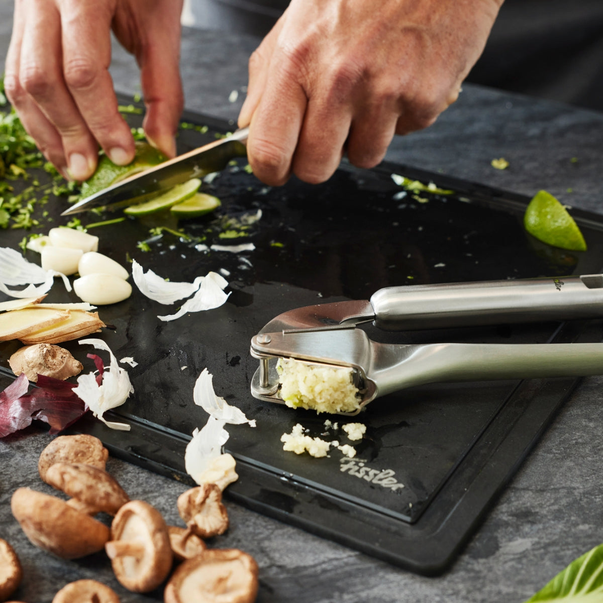 Fresh herbs and limes on a simple black board with garlic, mushrooms, ginger, and the Fissler Essential® garlic press.