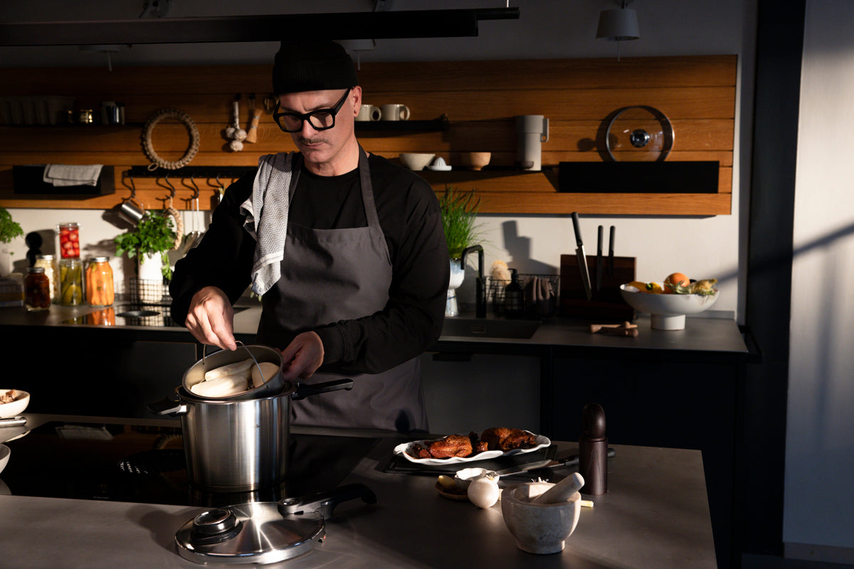 In einer sonnendurchfluteten, modernen Küche rührt eine Person mit Brille und Mütze gekonnt in einem Topf auf dem Herd, inspiriert von frischen Schnellkochtopf-Rezepten. Daneben liegen farbenfrohe Zutaten und Kräuter auf der Arbeitsplatte neben dem Fissler Vitavit® Premium 2-tlg. Set mit SKT-Buch – seit 1845 ein Aushängeschild deutscher Handwerkskunst –, bereit, Präzision beim Kochen, Langlebigkeit und zeitloses Design für eine weitere Gourmet-Kreation zu bieten.