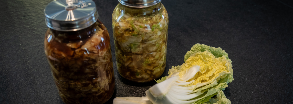 Zwei mit hausgemachtem Kimchi gefüllte Gläser stehen auf einer dunklen Arbeitsplatte neben einem halbierten Kopf frischem Chinakohl und vermitteln einen Eindruck von Präzision und Frische. Diese Inszenierung unterstreicht die Bedeutung hochwertiger Küchenutensilien – wie beispielsweise des Edelstahl-Kochgeschirrs oder des Zubehörs von Fissler – bei der Zubereitung und Aufbewahrung köstlicher hausgemachter Gerichte.