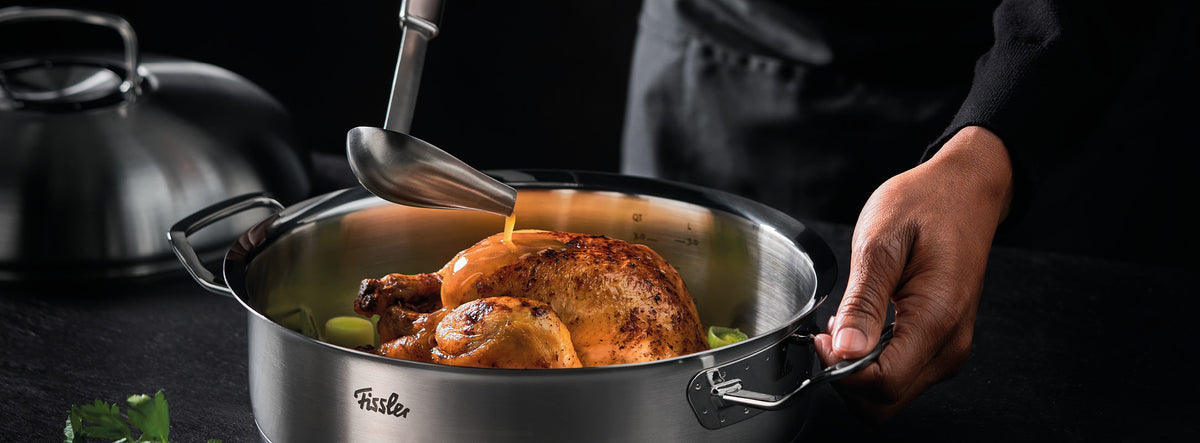 The sauce is poured over the roast chicken in a Fissler stainless steel pot, with herbs and lid nearby - an example of German quality cookware.