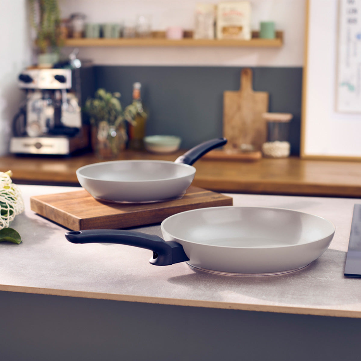 Experience German precision and timeless design with two Fissler Ceratal® Evo Classic pans with ergonomic black handles, one on a wooden cutting board. The ceramic nonstick coating ensures easy cooking and cleaning. In the background, discover