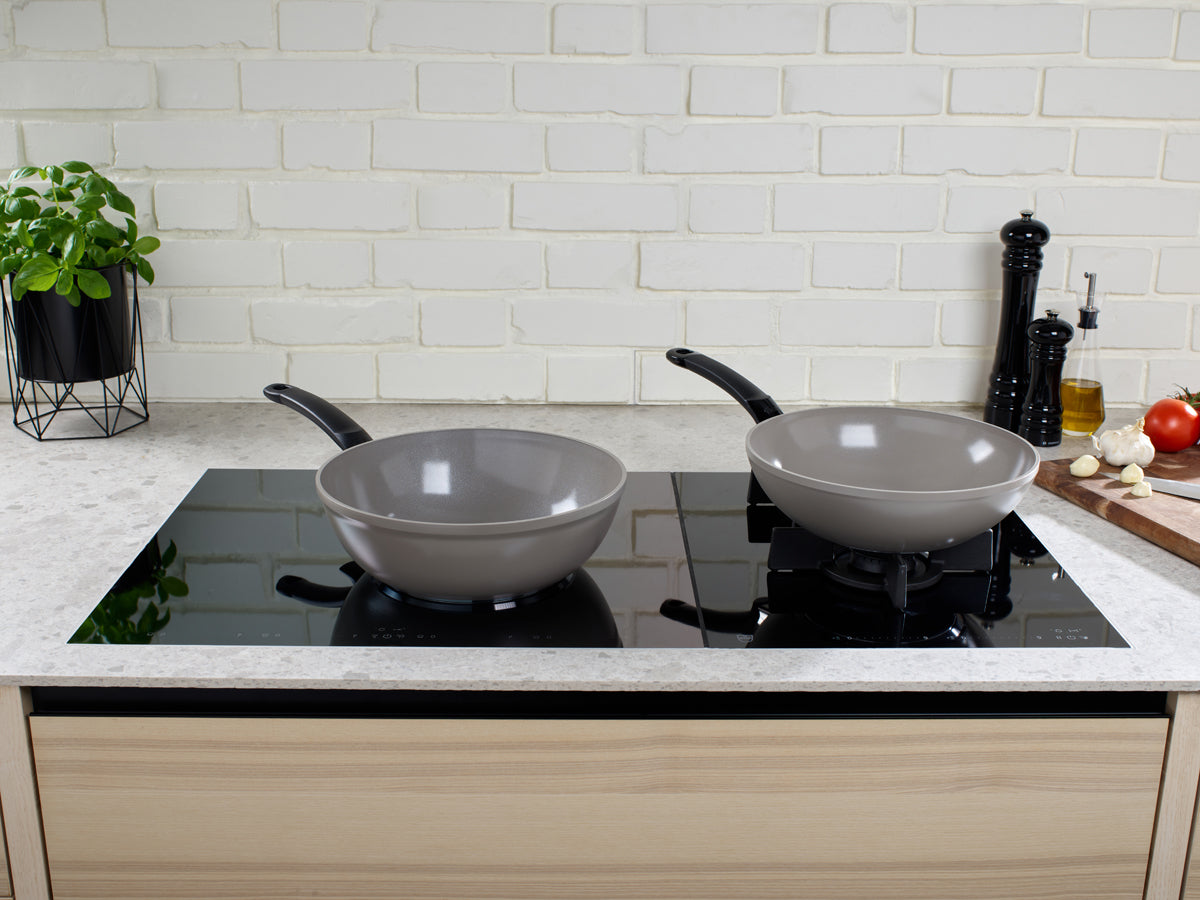Fissler Ceratal® wok pan and gray frying pan on a simple black stovetop, surrounded by fresh ingredients and a white brick backsplash.