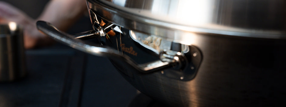 Close-up of a Fissler stainless steel pitcher with a precision-engineered handle reflecting light. Blurred hand and cup in the background.