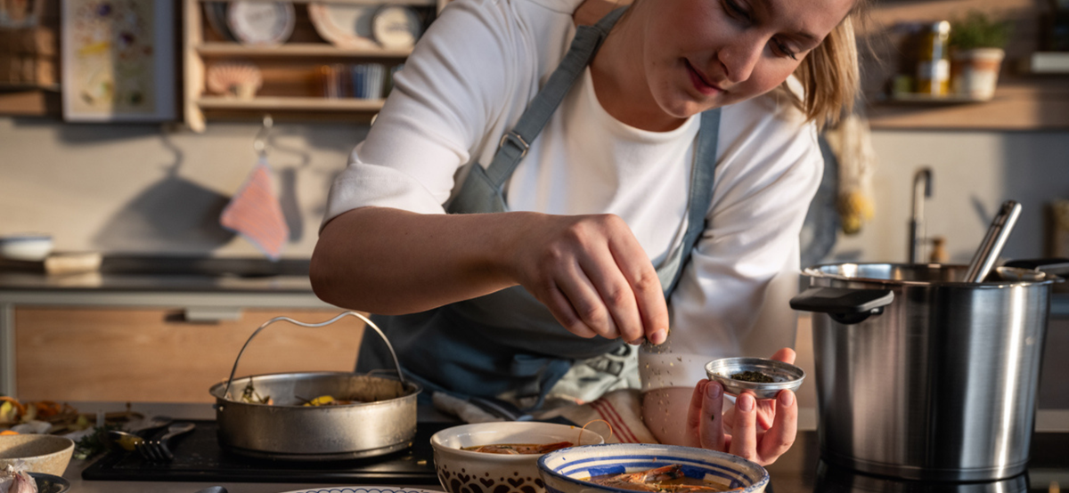Eine Person in einer Schürze bereitet das Essen zu und streut Kräuter in eine Schüssel, die von Fissler Kochgeschirr und Küchenzubehör umgeben ist.