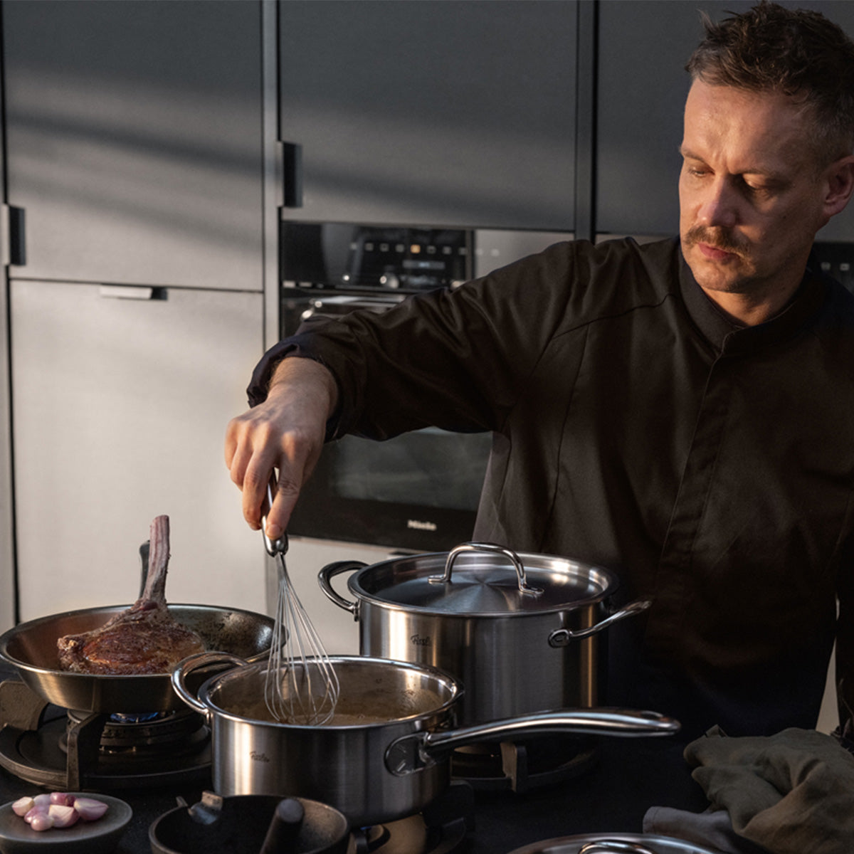 A man in a dark shirt cooks in a modern kitchen, stirring sauce in a Fissler M5 Pro-Ply® pot with metal lid. A steak sizzles in another Fissler pan from the same collection. Made from recycled stainless steel, induction-compatible and durable.