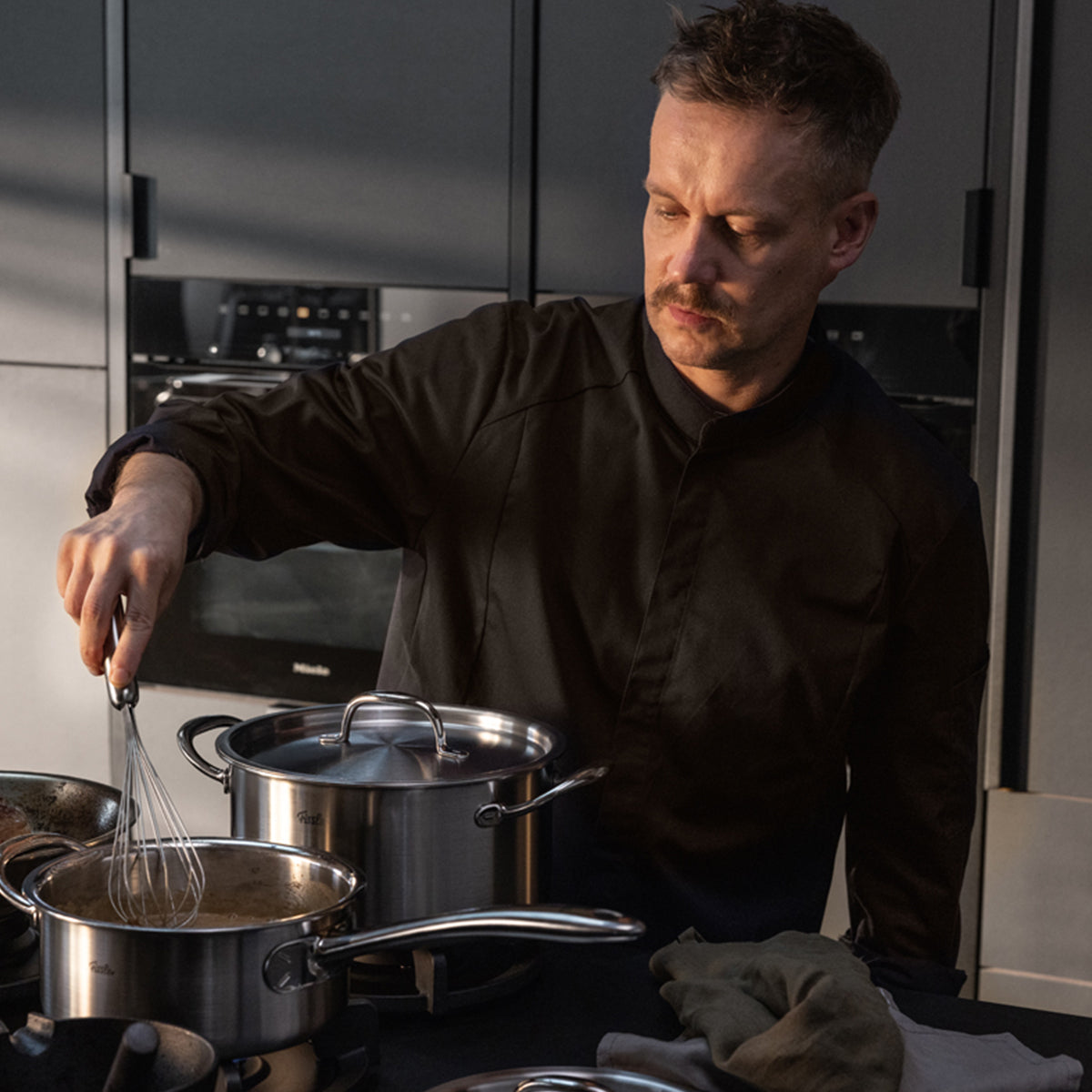 In a modern kitchen, a person in a dark chef's jacket stirs the Fissler M5 Pro-Ply® saucepan with metal lid, made from recycled stainless steel for induction use. Beside it stands another Fissler pot. Both reflect Fissler's 1845 tradition and are dishwasher-safe, making the