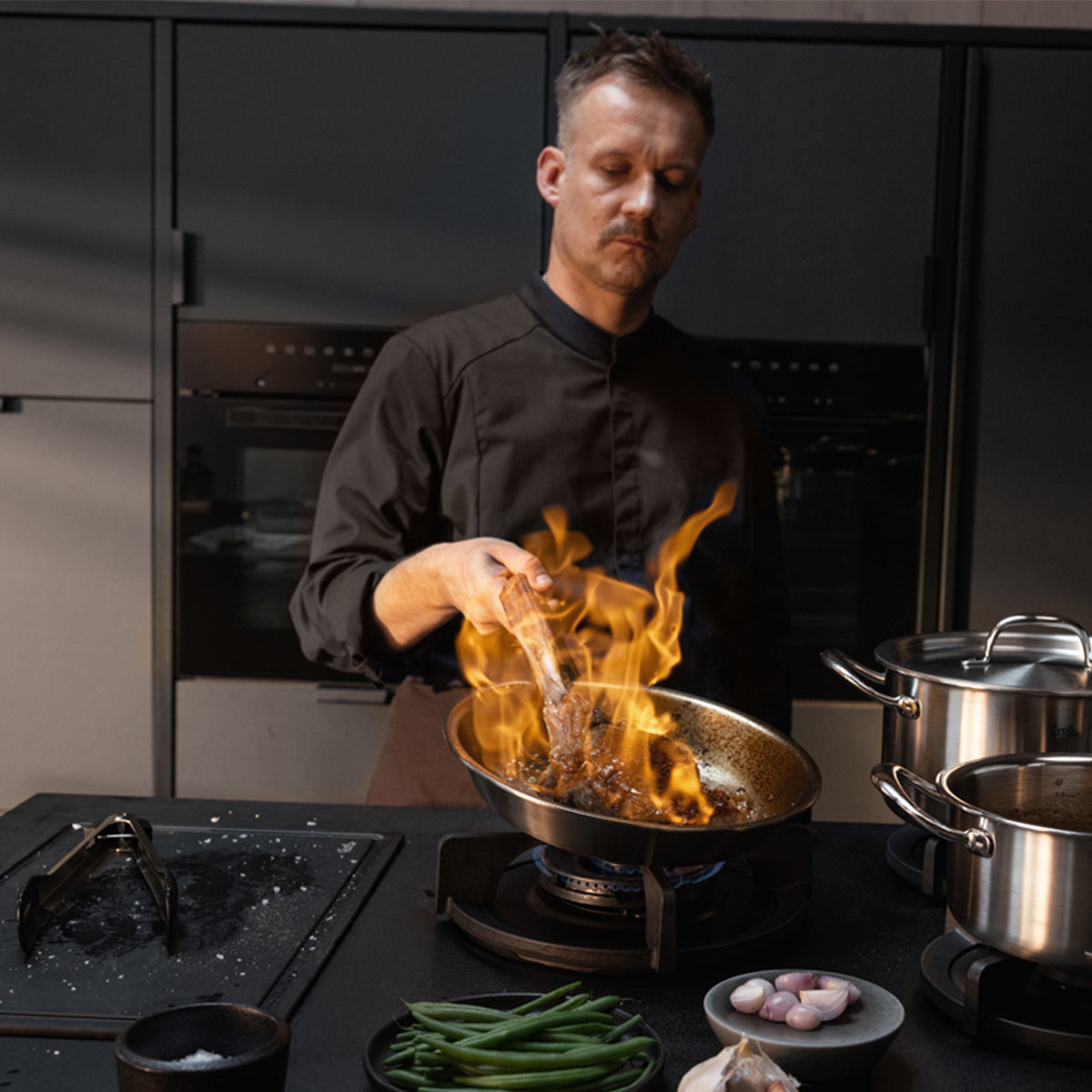Ein Koch in schwarzer Uniform brät gekonnt Fleisch in der Fissler M5 Pro-Ply®-Pfanne aus recyceltem Edelstahl, während die Flammen auf einem Gasherd elegant auflodern. Auf der Arbeitsplatte stehen hochwertige Fissler-Edelstahltöpfe neben frischen grünen Bohnen, Schalotten und Salz und spiegeln damit die Tradition der Marke wider: Präzisionstechnik und zeitloses deutsches Design. Alle gezeigten Kochgeschirrteile sind induktionsgeeignet und lassen sich mühelos reinigen – seit 1845 ein Markenzeichen für