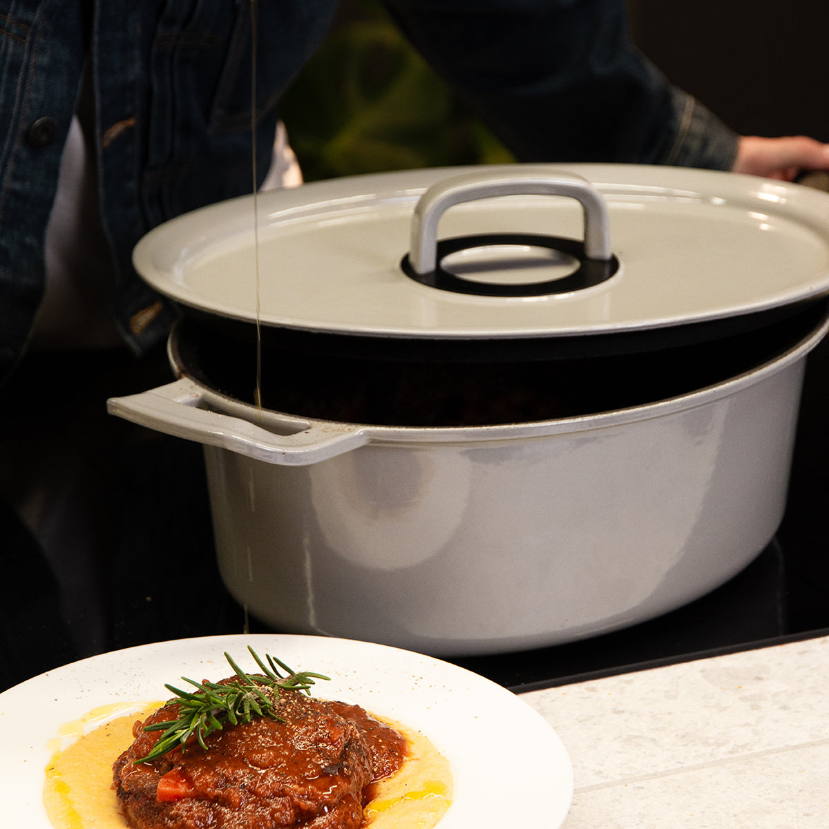 Öl wird über eine rosmarin-lackierte Schale gegossen, dahinter steht ein Fissler Gusseisen Bräter, der deutsche Präzision und Design zeigt.