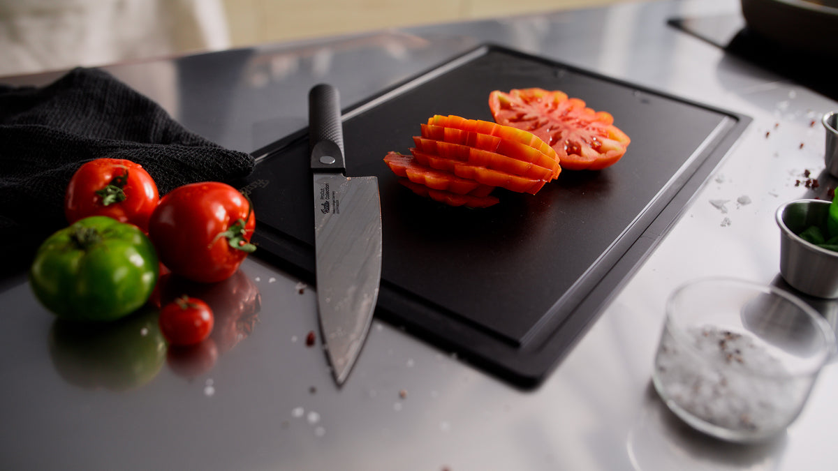 A refined kitchen scene showcases timeless Fissler quality: a black cutting board holds sliced tomatoes beside the Fissler ProCut Kochmesser 20 cm, crafted from extraharter Stahl for exceptional precision. Whole and green tomatoes, a towel, salt, and a glass complete the elegant arrangement on a sleek stainless steel countertop—reflecting Fissler’s heritage in German engineering and innovative cookware design since 1845.