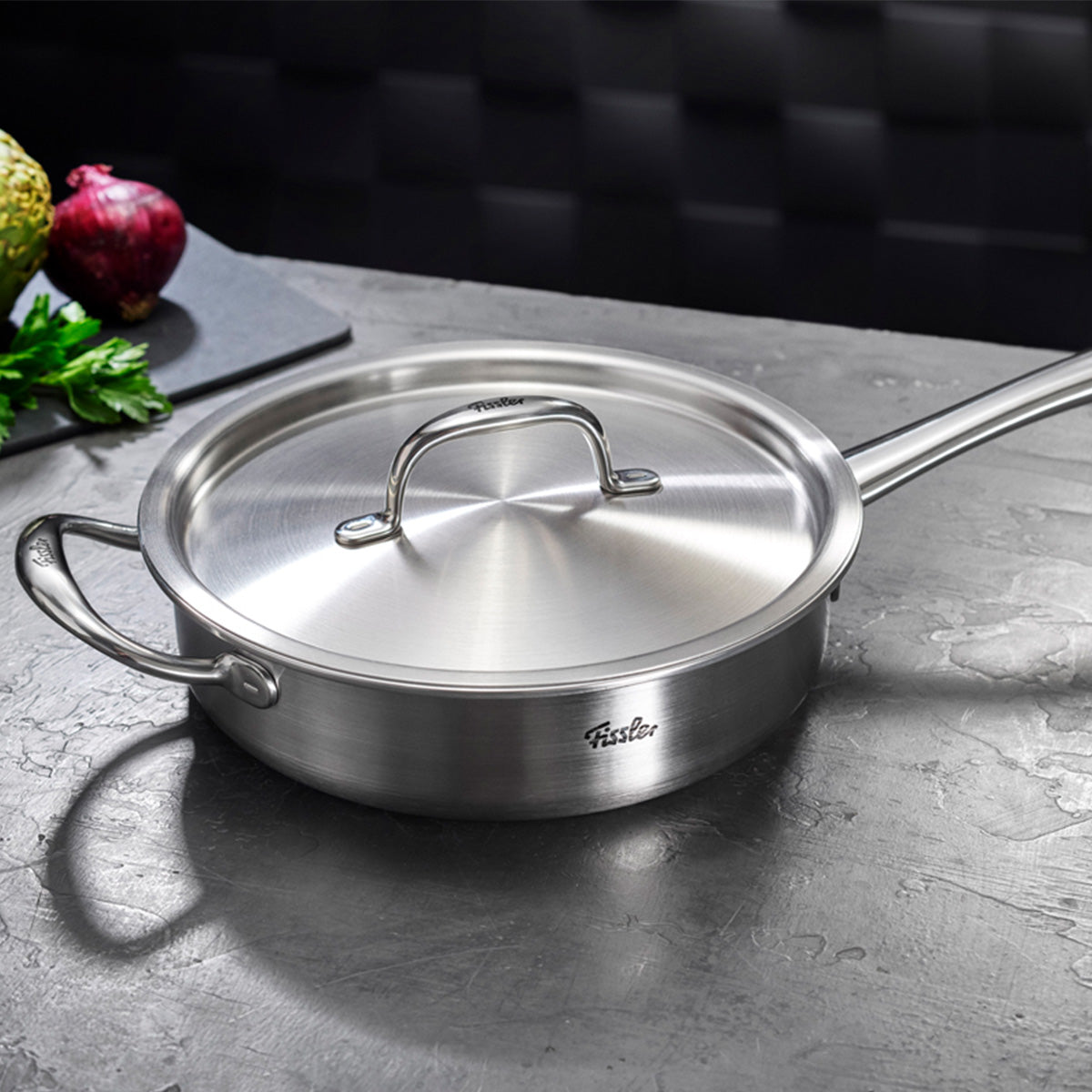 The 4-piece Fissler M5 Pro-Ply® set with metal lids sits on a grey worktop. Made in Germany from recycled stainless steel, the pots have sturdy handles and a brushed finish. In the background: onion, parsley and artichoke. This set stands for innovation,
