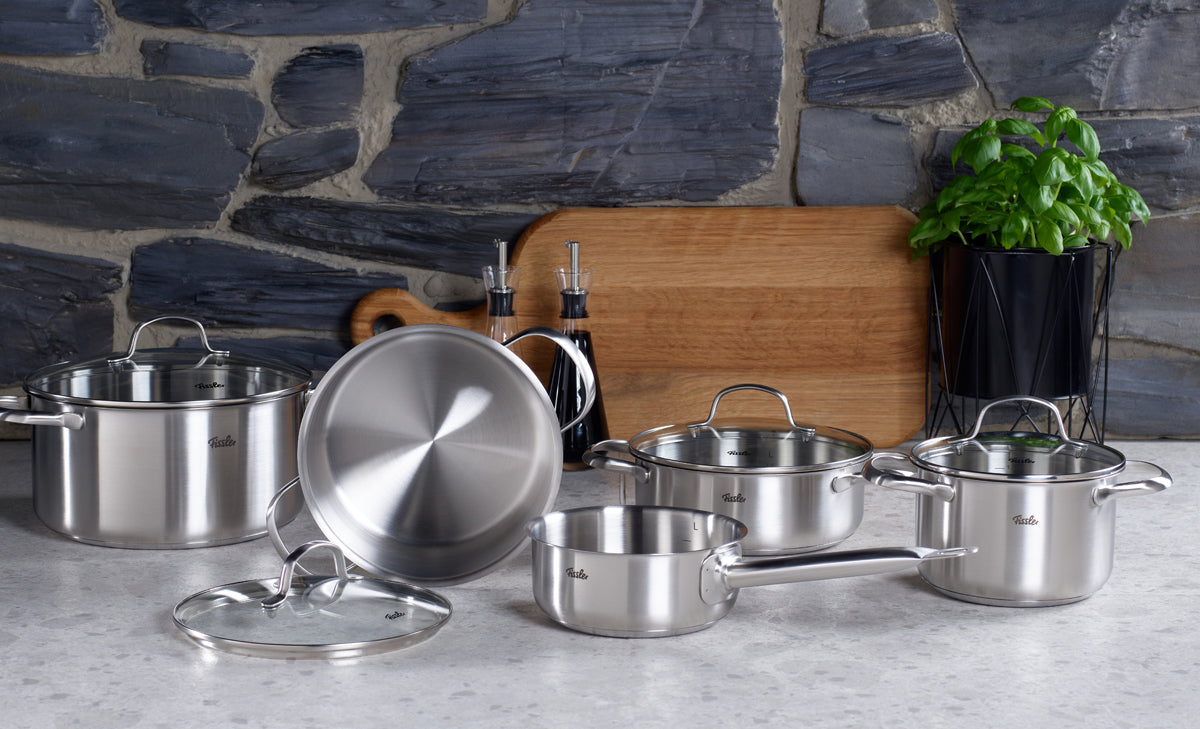 Ein Set aus Fissler San Francisco-Töpfen und -Pfannen aus Edelstahl mit Deckeln, das sich durch präzises deutsches Design auszeichnet und auf einer Küchenarbeitsplatte vor einer Steinwand angeordnet ist.