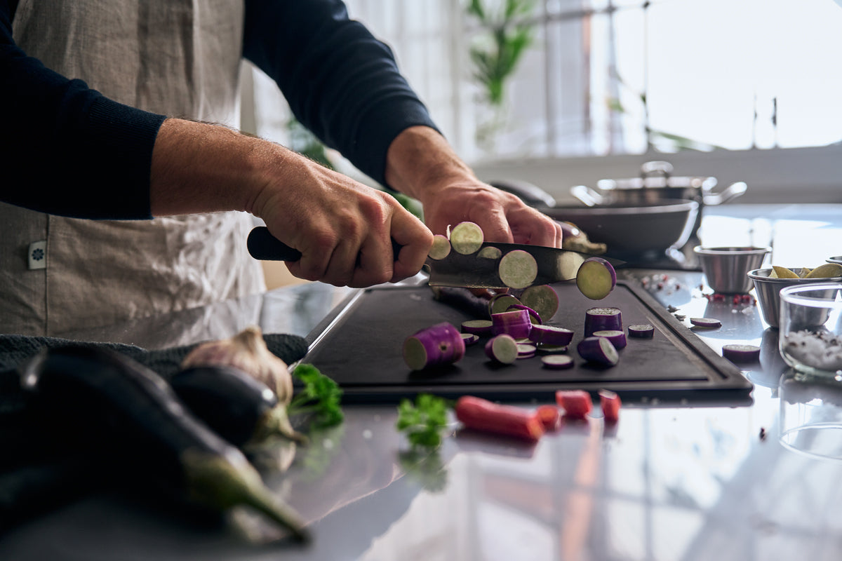A person in an apron skillfully slices purple eggplants with the precision-engineered Fissler ProCut Kochmesser 16 cm on a black cutting board, surrounded by fresh vegetables and premium Fissler stainless steel pots and kitchen accessories, showcasing German quality and timeless design in a modern kitchen setting.