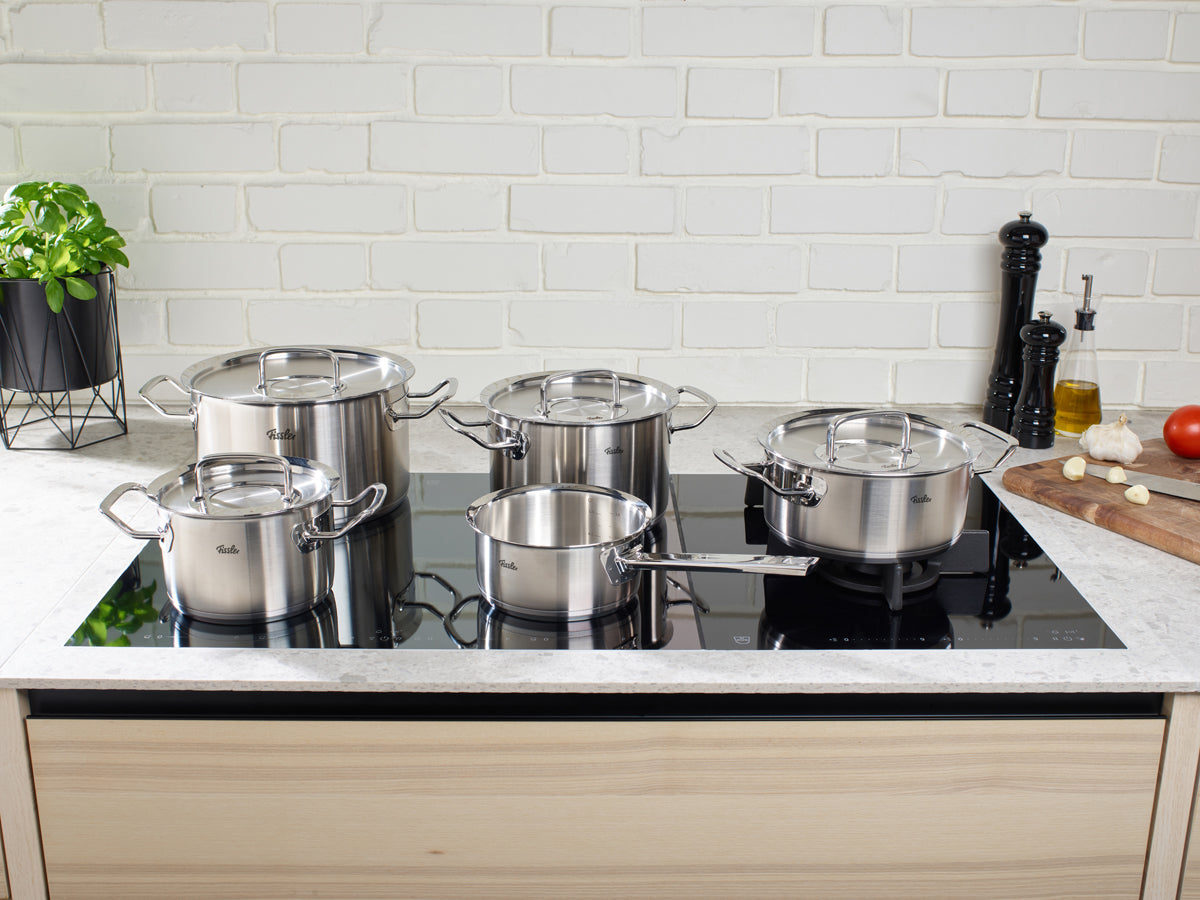 Five precision-engineered pots and pans from the Fissler Original-Profi Collection® 5-piece set—Kochtöpfe, Bratentopf, Stielkasserolle, and Servierpfanne—crafted from high-quality 18/10 stainless steel “Made in Germany,” are elegantly arranged on a modern black induction cooktop. To the right, a wooden board with sliced garlic, a tomato, and a pepper grinder highlights Fissler’s timeless commitment to innovation and durability against a classic white brick wall backdrop.