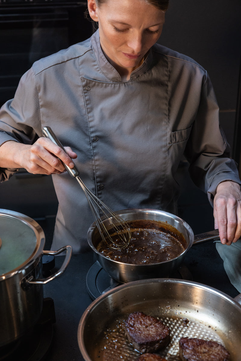 Ein Koch in grauer Uniform verrührt mit präzisen Handgriffen eine Soße in einer 20-cm-Sauteuse aus der Fissler Original-Profi Collection®, die unsere Tradition deutscher Ingenieurskunst widerspiegelt, auf einem Herd neben einer Pfanne mit angebratenen Steaks.