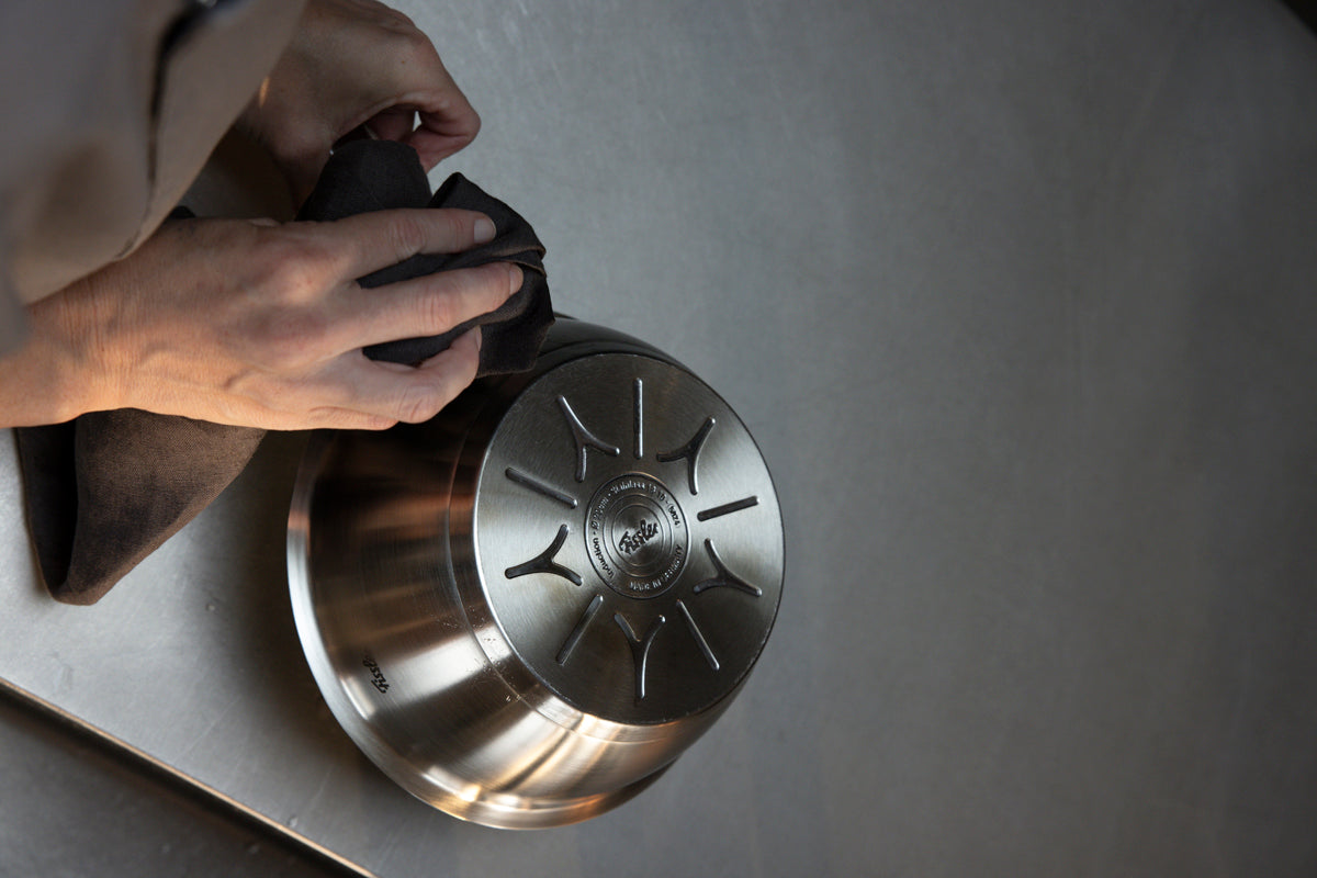 A person wipes the underside of a Fissler Original-Profi Collection® Sauteuse 20cm, reflecting Fissler’s heritage of precision engineering and durable design, with a cloth.