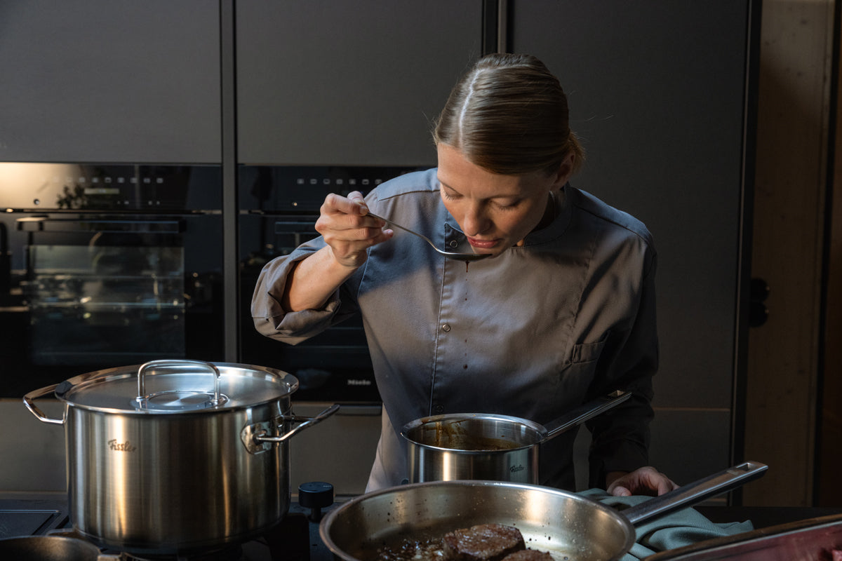 Ein Koch in grauer Uniform probiert auf einem modernen Küchenherd neben Töpfen aus der Fissler Original-Profi Collection® aus Edelstahl.