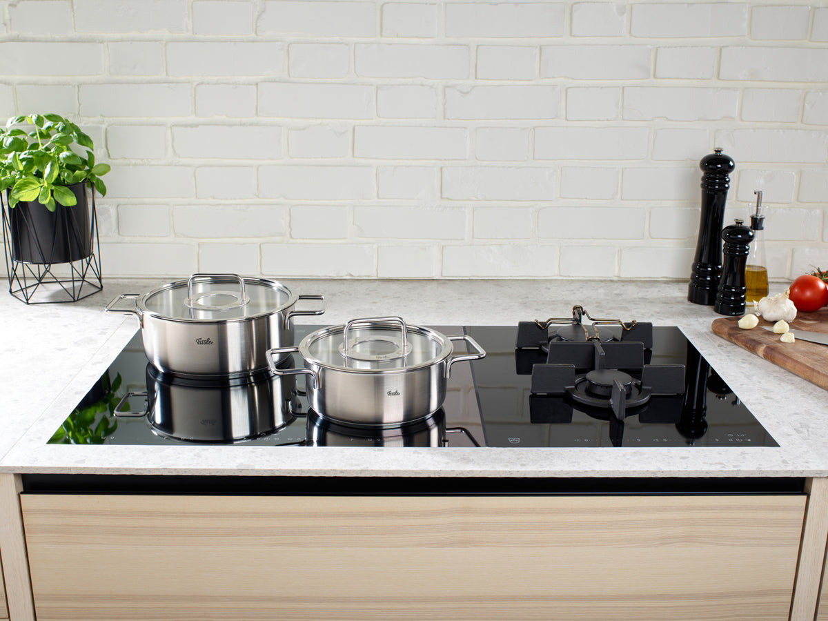 Eine moderne Küche mit dem 4-teiligen Topfset der Fissler Pure Collection® auf einem Glaskochfeld, frischen Zutaten und Kräutern in der Nähe.
