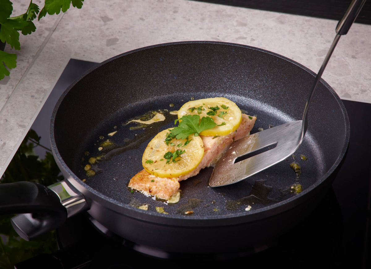 Salmon fillet with lemon and herbs in a Fissler Adamant® Comfort Frying Pan, lifted with a spatula; fresh parsley in the background.