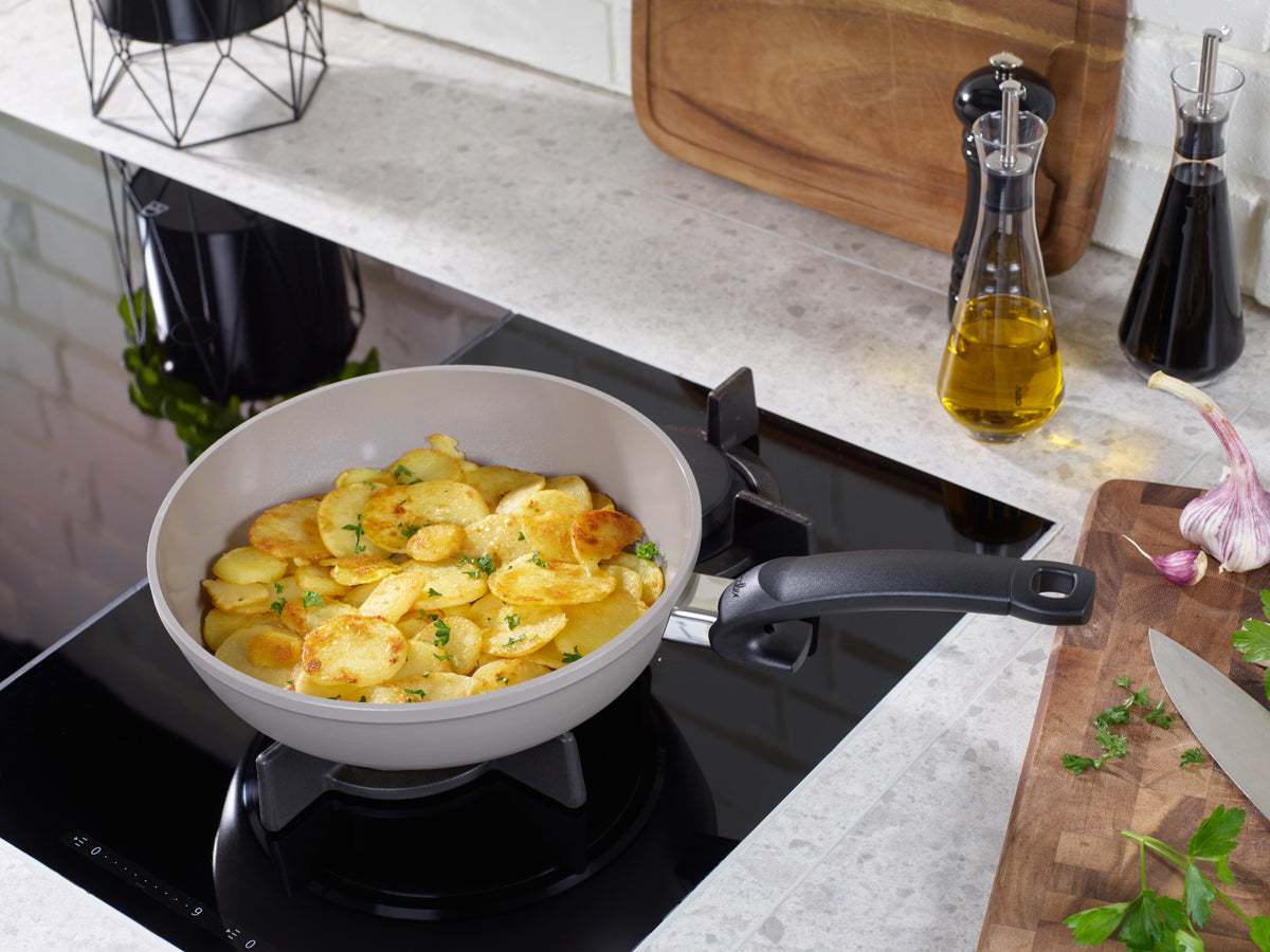 Fissler Ceratal® 2-pc. frying pan set cooking potatoes with herbs, German quality design, with oil, garlic, parsley on a light countertop.