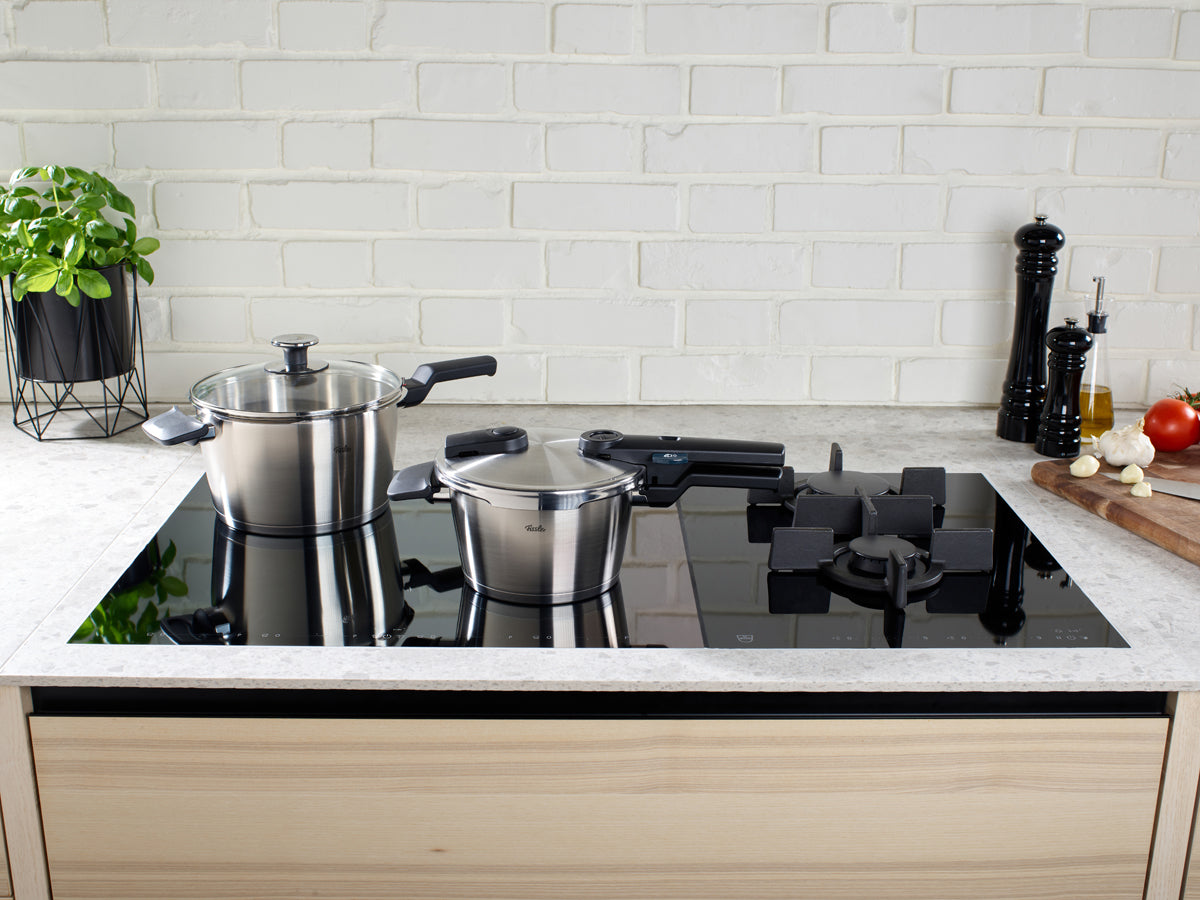 Fissler Vitaquick® Premium Pressure Cookers (2.5 & 4.5 L) on the stove; fresh vegetables, basil, oil on the counter - timeless German design.