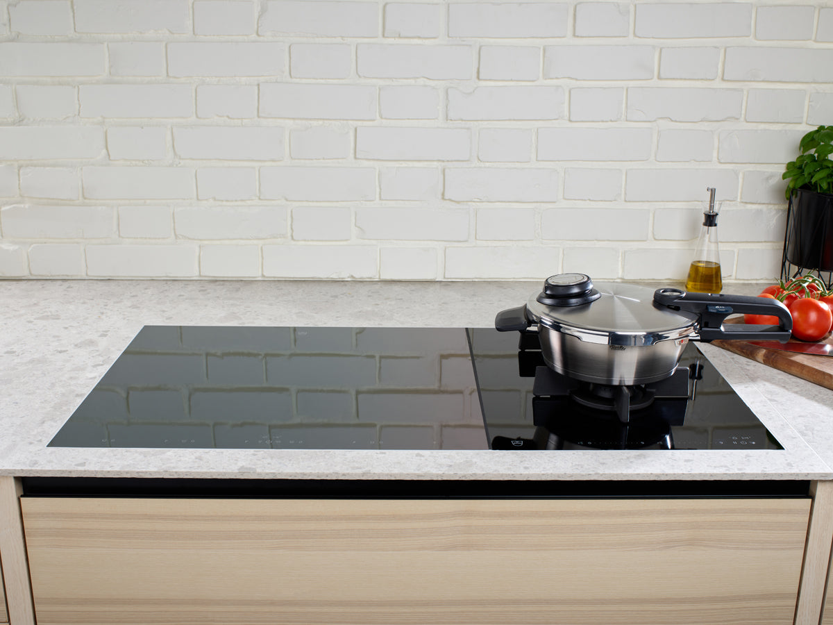 In a contemporary kitchen with a sleek black glass induction cooktop, the Fissler Vitavit® Premium Schnellbratpfanne crafted from high-quality Edelstahl 18/10 stainless steel sits elegantly with its matching lid on one burner. Signature touches such as cooking oil, ripe tomatoes, and fresh basil rest on the countertop against a classic white brick backsplash—an inviting scene showcasing Fissler’s German-made precision and timeless design.