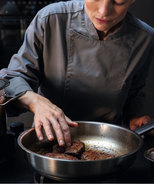 A chef in a gray uniform sears a steak in a Fissler stainless steel pan, showcasing German quality and innovation in a professional kitchen.