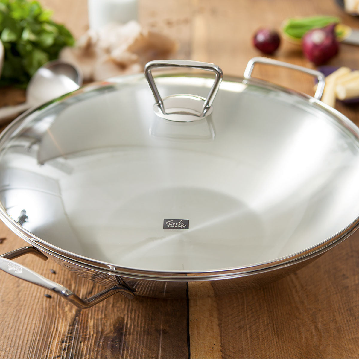 Fissler Kunming Wok, 36 cm, mit Glasdeckel, fachmännisch gefertigt mit präziser Technik und auf einer Holzunterlage stehend.