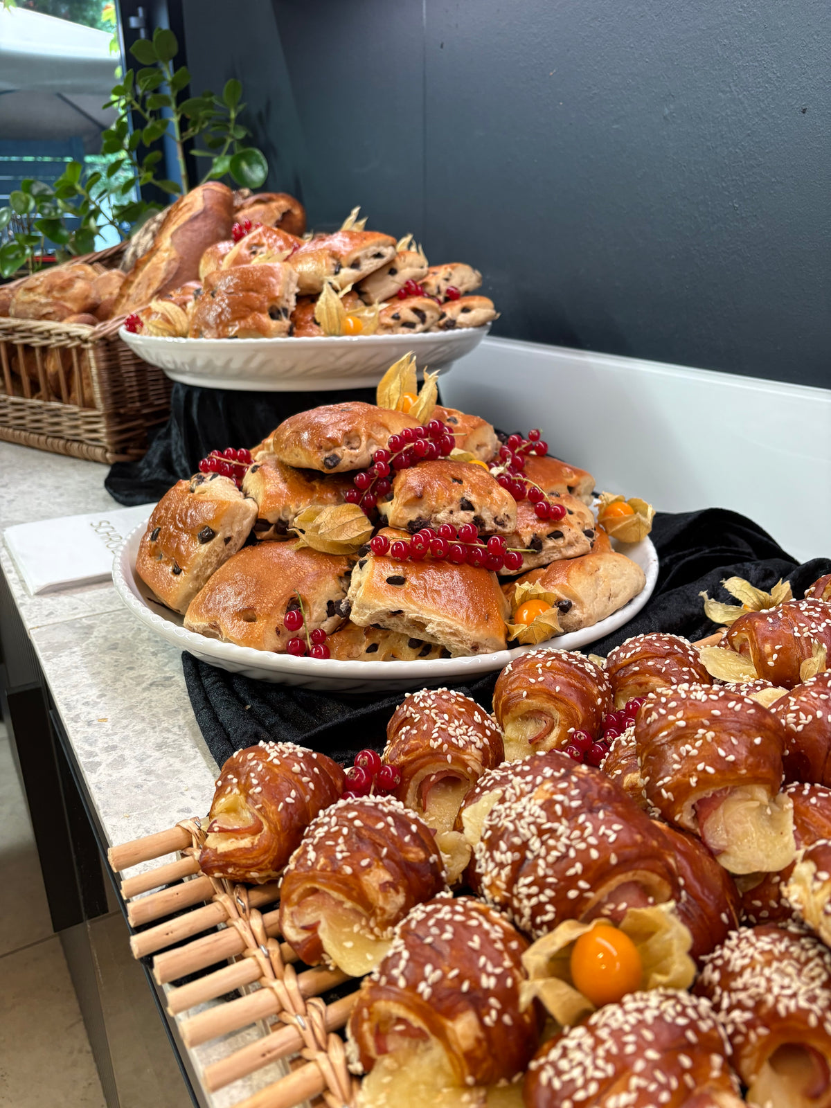 Verschiedene Backwaren wie Scones mit Schokoladenstückchen und Croissants mit Sesam, garniert mit Beeren und Kapstachelbeeren, elegant auf Platten und Tabletts auf einer Arbeitsplatte angerichtet, auf der das charakteristische Edelstahl-Serviergeschirr von Fissler präsentiert wird – ein Symbol für zeitlose deutsche Handwerkskunst und Kücheninnovation.
