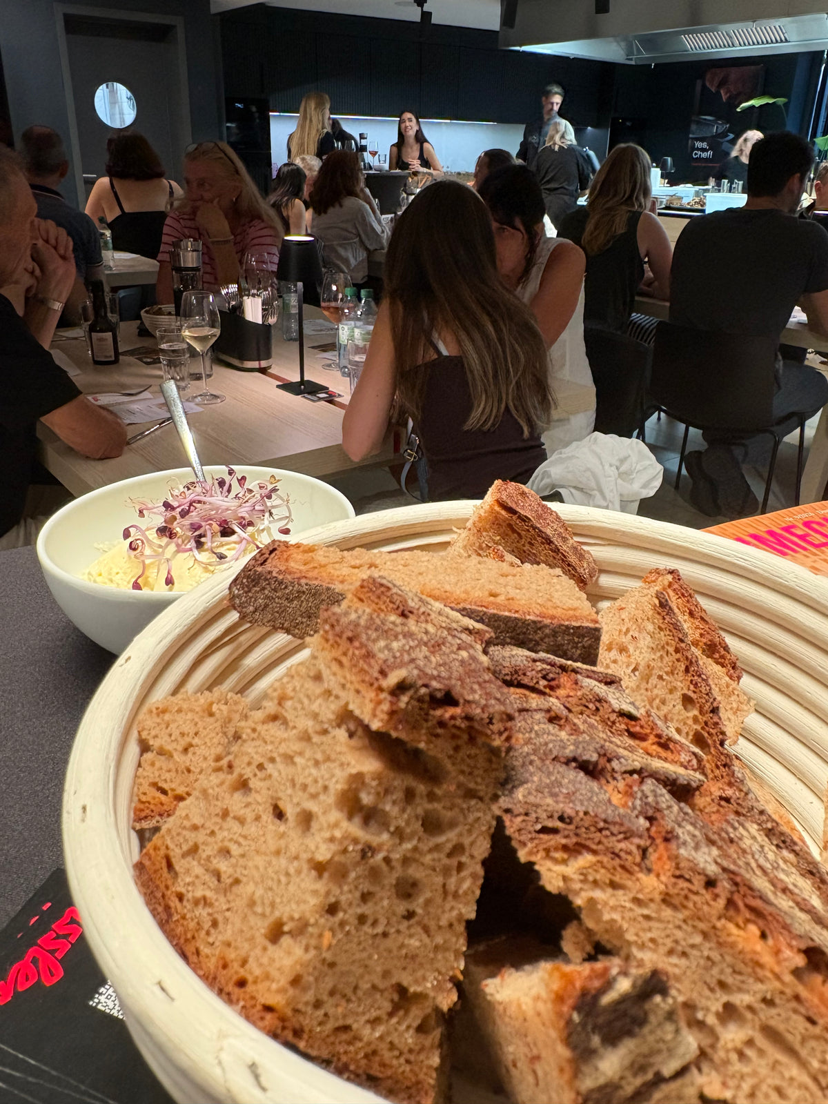 Eine Nahaufnahme eines Korbs voller Brotscheiben auf einem modernen Küchentisch, dazu ein frischer Salat in einer Fissler-Edelstahlschüssel. Im Hintergrund essen Menschen gemeinsam und unterhalten sich in einem einladenden Küchenbereich, der mit hochwertigem Fissler-Kochgeschirr ausgestattet ist – ein Beispiel für präzise Technik und zeitloses deutsches Design.