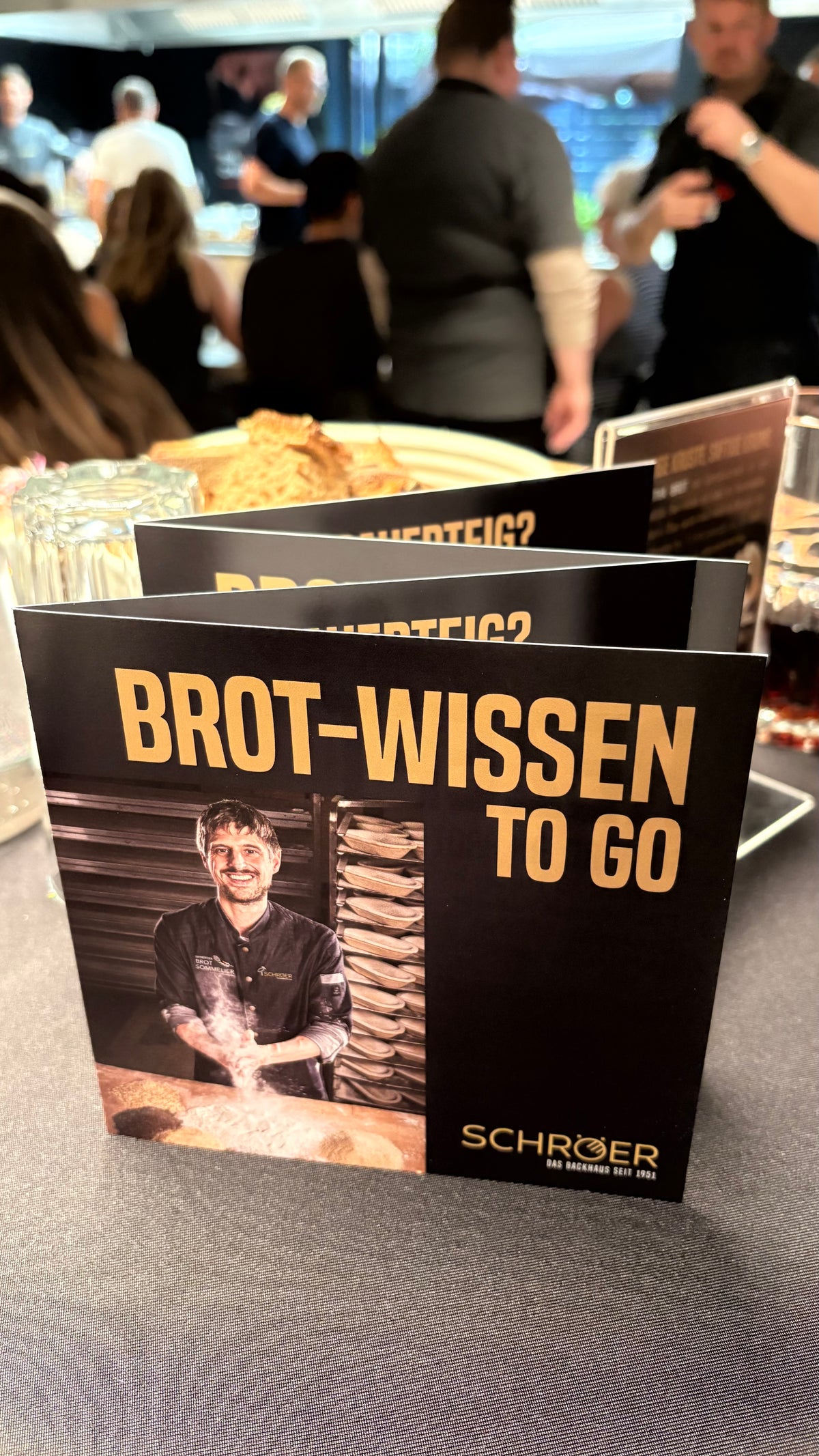 Eine Broschüre mit dem Titel „Brot-Wissen To Go“, auf deren Titelseite ein lächelnder Bäcker abgebildet ist, liegt elegant auf einem Tisch bei einer lebhaften Veranstaltung, umgeben von anderen Broschüren, edlem Glasgeschirr und den Teilnehmern im Hintergrund. Die Szene spiegelt Fisslers Bekenntnis zur deutschen Tradition und zur Innovation in der Kochkunst wider – eine einladende Atmosphäre, in der hochwertiges Kochgeschirr wie Edelstahltöpfe, Pfannen, Schnellkochtöpfe und Küchenzubehör das Ambiente