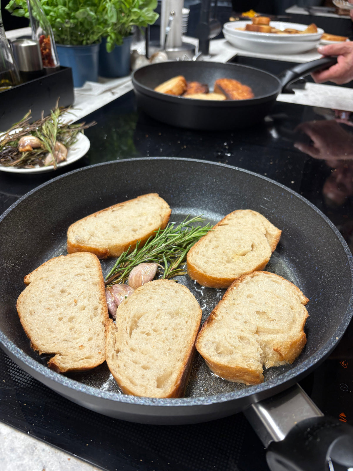 Fünf Scheiben Brot werden in einer präzise gefertigten Fissler-Pfanne mit frischem Rosmarin und Knoblauch perfekt geröstet, was die gleichmäßige Bräunung und die hervorragende Wärmeverteilung verdeutlicht. Im Hintergrund stehen eine weitere hochwertige Fissler-Pfanne mit knusprigem Brot sowie ein Teller mit duftenden Kräutern und Knoblauch auf einer modernen Küchenarbeitsplatte – ein Spiegelbild des Engagements der Marke für innovatives Design, Langlebigkeit und deutsche Handwerkskunst.