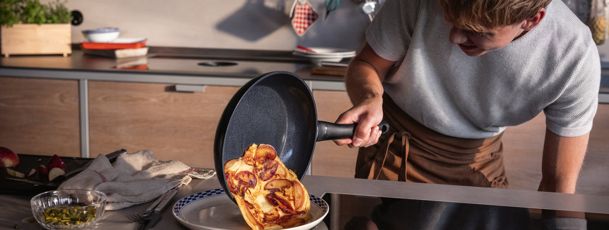 Eine Person schiebt ein goldenes Omelett aus einer Fissler-Pfanne auf einen Teller; hochwertiges Kochgeschirr und frische Zutaten stehen auf der Theke.