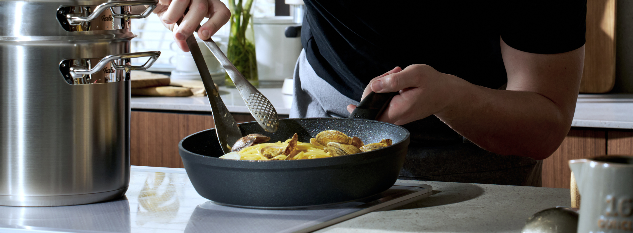 In einer modernen Küche bereitet eine Person auf einem eleganten Kochfeld in einer schwarzen Fissler-Pfanne mit einer Zange in Scheiben geschnittenes Gemüse zu, daneben steht ein charakteristischer Fissler-Edelstahltopf. Zu sehen sind nur der Oberkörper und die Arme der Person, wodurch die Verwendung von hochwertigem, in Deutschland entwickeltem Kochgeschirr hervorgehoben wird, das für Qualität und Innovation bekannt ist.