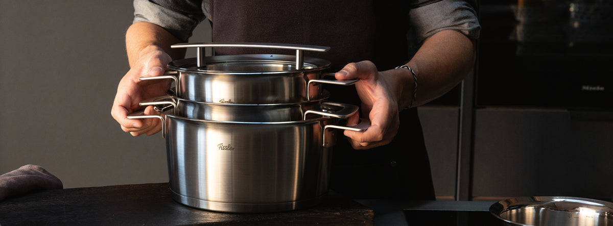 Eine Person stapelt die in Deutschland hergestellten Fissler-Edelstahltöpfe mit ihren haltbaren Griffen und der zeitlosen, polierten Oberfläche auf einer Küchentheke.