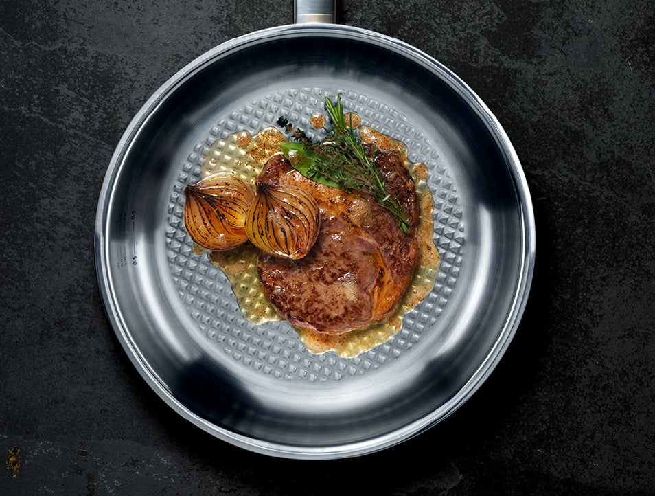 A perfectly fried steak with onions, rosemary, and sauce in a Fissler stainless steel pan on a dark countertop.