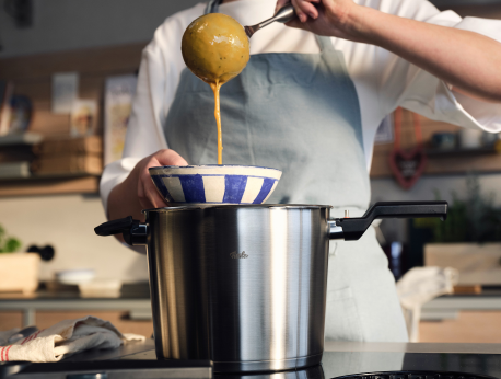 In einer modernen Küche schöpft eine Person in einer Schürze Suppe aus einem hochwertigen Fissler-Edelstahltopf in eine blau-weiß gestreifte Schüssel und präsentiert damit präzisionsgefertigtes Kochgeschirr, das für seine Langlebigkeit und sein zeitloses deutsches Design bekannt ist.