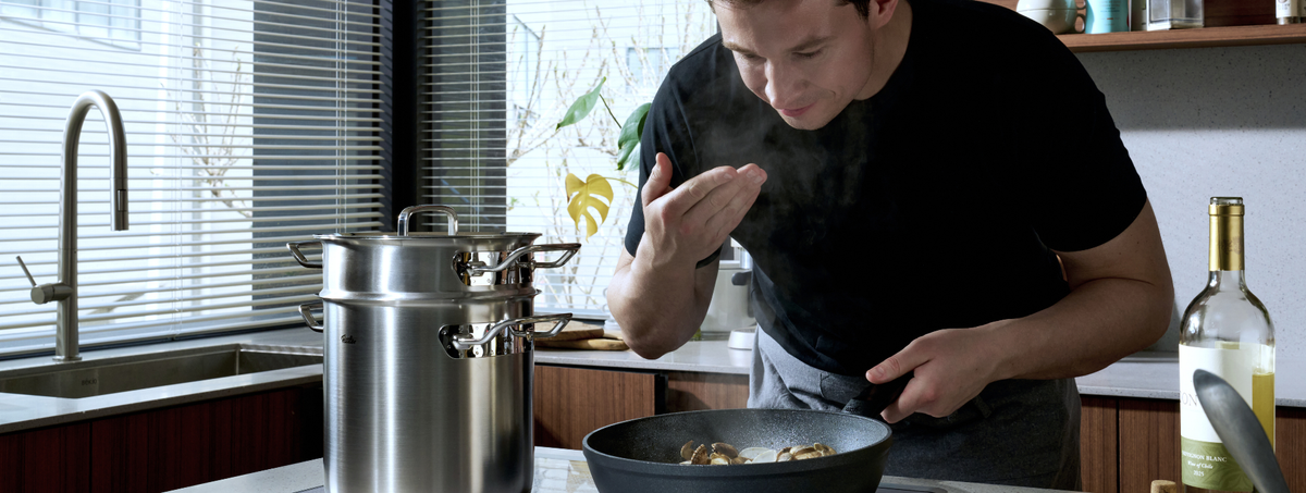 In einer sonnendurchfluteten Küche probiert ein Hobbykoch ein Gericht aus einer hochwertigen Fissler-Pfanne. Ein glänzender Fissler-Edelstahltopf steht nebenan auf dem Herd, während eine Flasche Weißwein auf der Arbeitsplatte steht. Präzisionsgefertigtes Kochgeschirr und elegantes deutsches Design runden den einladenden Kochbereich ab, während natürliches Licht durch die Jalousien hereinströmt.