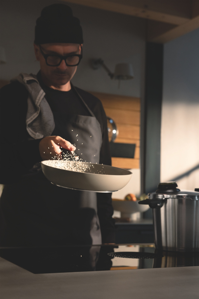 In einer sonnendurchfluteten, modernen Küche schwenkt eine Person mit Brille und Schürze gekonnt Reis in einer Edelstahlpfanne von Fissler und unterstreicht damit die Tradition der Marke in Sachen Präzisionstechnik und zeitloses deutsches Design.