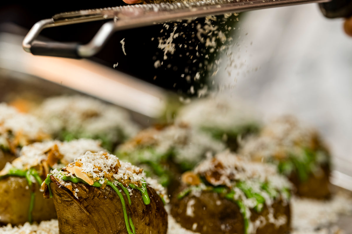 Nahaufnahme einer Hand, die mit einer präzisionsgefertigten Fissler-Reibe frischen Käse über goldbraun gebratene Kartoffeln reibt, die elegant mit leuchtend grünen Kräutern und herzhaften Pilzen garniert sind. Feine Käsespäne fallen sanft auf das Gericht und unterstreichen die Qualität und Innovation der Fissler-Küchenaccessoires.